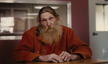 Movie still from “The Mustang” (2019), directed by Laure de Clermont-Tonnerre – An old man with a long beard sitting at a table; Medium shot, Over the shoulder angle
