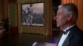 Movie still from “Naked Gun 33 1/3: The Final Insult” (1994), directed by Peter Segal – An older man in a suit is talking into a microphone; Medium shot, Over the shoulder angle