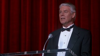 Movie still from “Naked Gun 33 1/3: The Final Insult” (1994), directed by Peter Segal – An older man in a tuxedo is speaking at a podium; Medium shot, Low angle