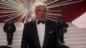 Movie still from “Naked Gun 33 1/3: The Final Insult” (1994), directed by Peter Segal – A man in a suit and bow tie standing in front of stairs; Medium shot, Low angle
