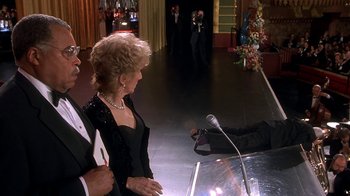 Movie still from “Naked Gun 33 1/3: The Final Insult” (1994), directed by Peter Segal – A woman standing at a podium in front of a microphone; Wide shot, High angle