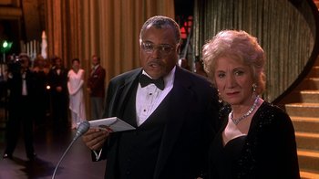 Movie still from “Naked Gun 33 1/3: The Final Insult” (1994), directed by Peter Segal – An older man and woman in formal wear holding a microphone; Medium shot, High angle