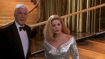 Movie still from “Naked Gun 33 1/3: The Final Insult” (1994), directed by Peter Segal – A woman in a silver dress standing next to a man in a tuxedo; Medium shot, Over the shoulder angle