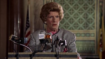 Movie still from “The Naked Gun: From the Files of Police Squad!” (1988), directed by David Zucker – An older woman standing at a podium with microphones; Close Up shot, Low angle