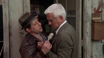 Movie still from “The Naked Gun: From the Files of Police Squad!” (1988), directed by David Zucker – A man in a suit and a hat is holding a tie around a man's neck; Close Up shot, Over the shoulder angle