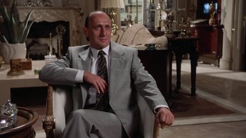 Movie still from “The Naked Gun: From the Files of Police Squad!” (1988), directed by David Zucker – A man in a suit and tie sitting in a chair in front of a fire place; Medium shot, Over the shoulder angle