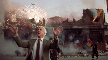 Movie still from “The Naked Gun: From the Files of Police Squad!” (1988), directed by David Zucker – An older man in a suit and tie standing in front of fireworks; Medium shot, Low angle