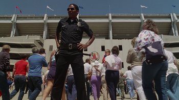 Movie still from “The Naked Gun: From the Files of Police Squad!” (1988), directed by David Zucker – A man in a police uniform standing in front of a group of onlookers; Wide shot, Low angle
