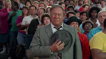 Movie still from “The Naked Gun: From the Files of Police Squad!” (1988), directed by David Zucker – A man in a suit holding a hat in front of a crowd of onlookers; Close Up shot, Over the shoulder angle