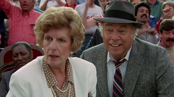 Movie still from “The Naked Gun: From the Files of Police Squad!” (1988), directed by David Zucker – An older man and a woman are sitting in front of a crowd; Close Up shot, Over the shoulder angle