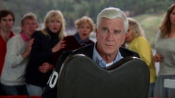 Movie still from “The Naked Gun: From the Files of Police Squad!” (1988), directed by David Zucker – An older man is looking into the camera while a group of people watch; Medium shot, Over the shoulder angle