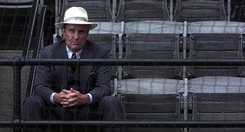 Movie still from “The Natural” (1984), directed by Barry Levinson – An older man in a suit and hat sitting in front of a chair; Medium shot, Low angle
