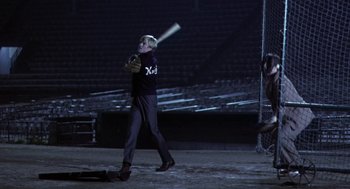 Movie still from “The Natural” (1984), directed by Barry Levinson – A man swinging a baseball bat on a baseball field at night; Wide shot, Over the shoulder angle