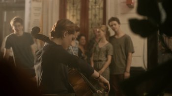 Movie still from “Charlie Countryman” (2013), directed by Fredrik Bond – A group of people standing in a room with a woman playing a cello; Wide shot, Over the shoulder angle