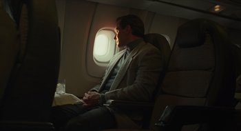 Movie still from “The Nest” (2020), directed by Sean Durkin – A man sitting in a chair on an airplane looking out the window; Medium shot, Low angle