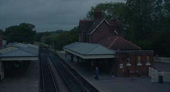 Movie still from “The Nest” (2020), directed by Sean Durkin – A man standing on the side of a train track next to a train station; Extreme Wide shot, High angle