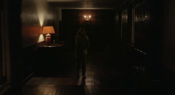 Movie still from “The Nest” (2020), directed by Sean Durkin – A woman standing in a dark room in front of a lamp; Wide shot, Low angle