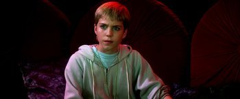 Movie still from “The NeverEnding Story II: The Next Chapter” (1990), directed by George Miller – A young man with blonde hair sitting in front of a red background; Close Up shot, High angle