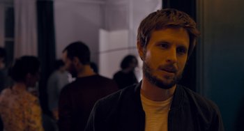 Movie still from “The Night Eats the World” (2018), directed by Dominique Rocher – A man with a beard is standing in front of a group of people; Close Up shot, Over the shoulder angle