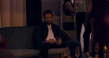 Movie still from “The Night Eats the World” (2018), directed by Dominique Rocher – A man sitting on top of a couch holding a drink; Medium shot, Over the shoulder angle