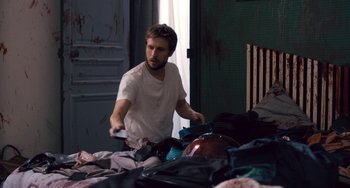 Movie still from “The Night Eats the World” (2018), directed by Dominique Rocher – A man standing on a bed in front of a pile of luggage; Medium shot, Over the shoulder angle