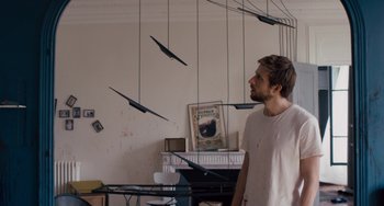 Movie still from “The Night Eats the World” (2018), directed by Dominique Rocher – A man standing in a room next to a fireplace; Medium shot, High angle