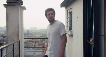Movie still from “The Night Eats the World” (2018), directed by Dominique Rocher – A man standing on a balcony with a city view in the background; Medium shot, Low angle