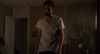 Movie still from “The Night Eats the World” (2018), directed by Dominique Rocher – A man standing in a dark room wearing a t - shirt; Medium shot, Low angle