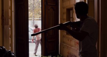 Movie still from “The Night Eats the World” (2018), directed by Dominique Rocher – A man holding a rifle in front of an open door; Medium shot, Over the shoulder angle