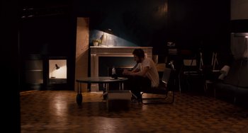 Movie still from “The Night Eats the World” (2018), directed by Dominique Rocher – A man sitting on a chair in front of a fireplace; Wide shot, High angle
