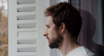 Movie still from “The Night Eats the World” (2018), directed by Dominique Rocher – A man with a beard looking out of a window; Close Up shot, Low angle
