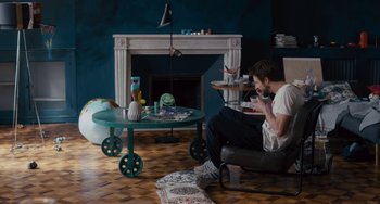 Movie still from “The Night Eats the World” (2018), directed by Dominique Rocher – A man sitting in a chair in front of a fireplace; Wide shot, High angle