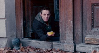 Movie still from “The Night Eats the World” (2018), directed by Dominique Rocher – A young man holding a bowl of food looking out of a window; Medium shot, Low angle
