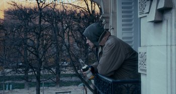 Movie still from “The Night Eats the World” (2018), directed by Dominique Rocher – A man sitting on a balcony holding a bottle of beer; Medium shot, Over the shoulder angle
