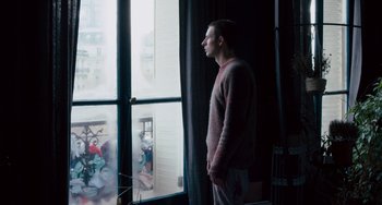 Movie still from “The Night Eats the World” (2018), directed by Dominique Rocher – A man standing in front of a window looking outside; Medium shot, Low angle