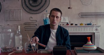 Movie still from “The Night Eats the World” (2018), directed by Dominique Rocher – A man sitting at a table with a pencil and pencil sharpener in front of him; Medium shot, Low angle