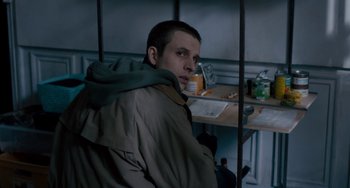 Movie still from “The Night Eats the World” (2018), directed by Dominique Rocher – A man sitting in front of a table with a bottle; Medium shot, High angle