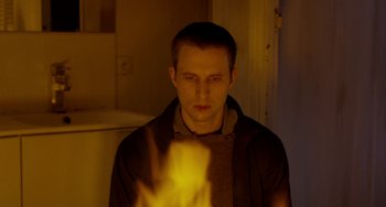 Movie still from “The Night Eats the World” (2018), directed by Dominique Rocher – A young man in a dark room with a fire in his hand; Close Up shot, High angle