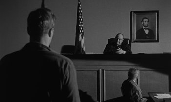 Movie still from “The Night of the Hunter” (1955), directed by Charles Laughton – A man sitting in front of an american flag in a courtroom; Wide shot, Over the shoulder angle