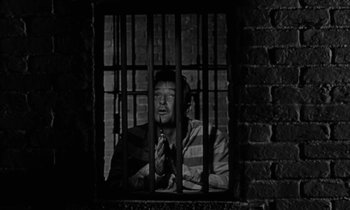 Movie still from “The Night of the Hunter” (1955), directed by Charles Laughton – A man sitting in front of a jail cell with his hands clasped behind bars; Medium shot, Low angle
