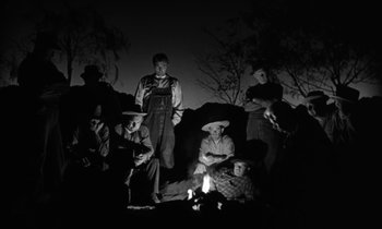 Movie still from “The Night of the Hunter” (1955), directed by Charles Laughton – A black and white photo of a group of people sitting around a fire; Wide shot, High angle