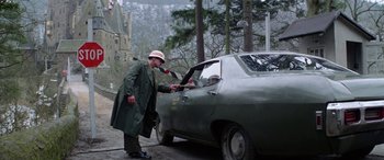 Movie still from “The Ninth Configuration” (1980), directed by William Peter Blatty – A man in a green raincoat and a white hat is standing next to a car; Wide shot, High angle