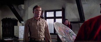 Movie still from “The Ninth Configuration” (1980), directed by William Peter Blatty – A man standing in front of a painting in front of another man; Medium shot, Low angle