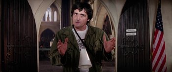 Movie still from “The Ninth Configuration” (1980), directed by William Peter Blatty – A man in a green jacket is holding his hands up; Medium shot, Low angle