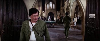Movie still from “The Ninth Configuration” (1980), directed by William Peter Blatty – A man walking in a large room with people in the background; Medium shot, Over the shoulder angle