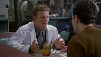 Movie still from “The Object of My Affection” (1998), directed by Nicholas Hytner – A man in a white lab coat sitting at a table with a drink; Medium shot, Over the shoulder angle