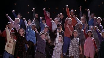 Movie still from “The Object of My Affection” (1998), directed by Nicholas Hytner – A large group of children standing on a stage with their hands in the air; Medium shot, High angle