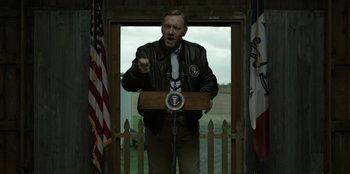 Movie still from “House of Cards” (2013), created by Beau Willimon – A man in a brown jacket is speaking at a podium; Medium shot, Over the shoulder angle