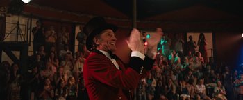 Movie still from “The One and Only Ivan” (2020), directed by Thea Sharrock – A man wearing a red jacket and a black top hat clapping in front of a crowd; Medium shot, Over the shoulder angle