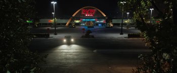 Movie still from “The One and Only Ivan” (2020), directed by Thea Sharrock – A car driving down a street at night past a big top mall; Extreme Wide shot, High angle